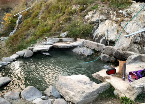 A natural hot spring with stone edges, surrounded by rocks and grass, featuring a small water source and a backpack nearby.